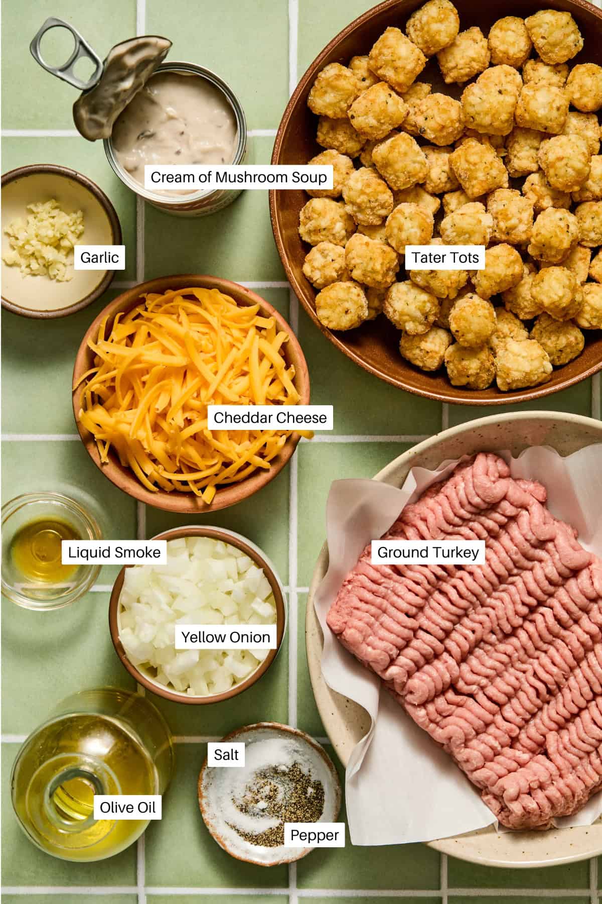 Ingredients for a Turkey Tater Tot Casserole are arranged in bowls: tater tots, ground turkey, shredded cheddar cheese, diced onion, minced garlic, olive oil, cream of mushroom soup, liquid smoke, salt, and pepper on a green-tiled surface.