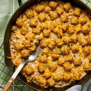 A cast iron skillet filled with a hearty Turkey Tater Tot Casserole, topped with melted cheese and chopped chives. A serving spoon rests in the dish, and a checkered cloth is nearby. The skillet sits on a cooling rack.