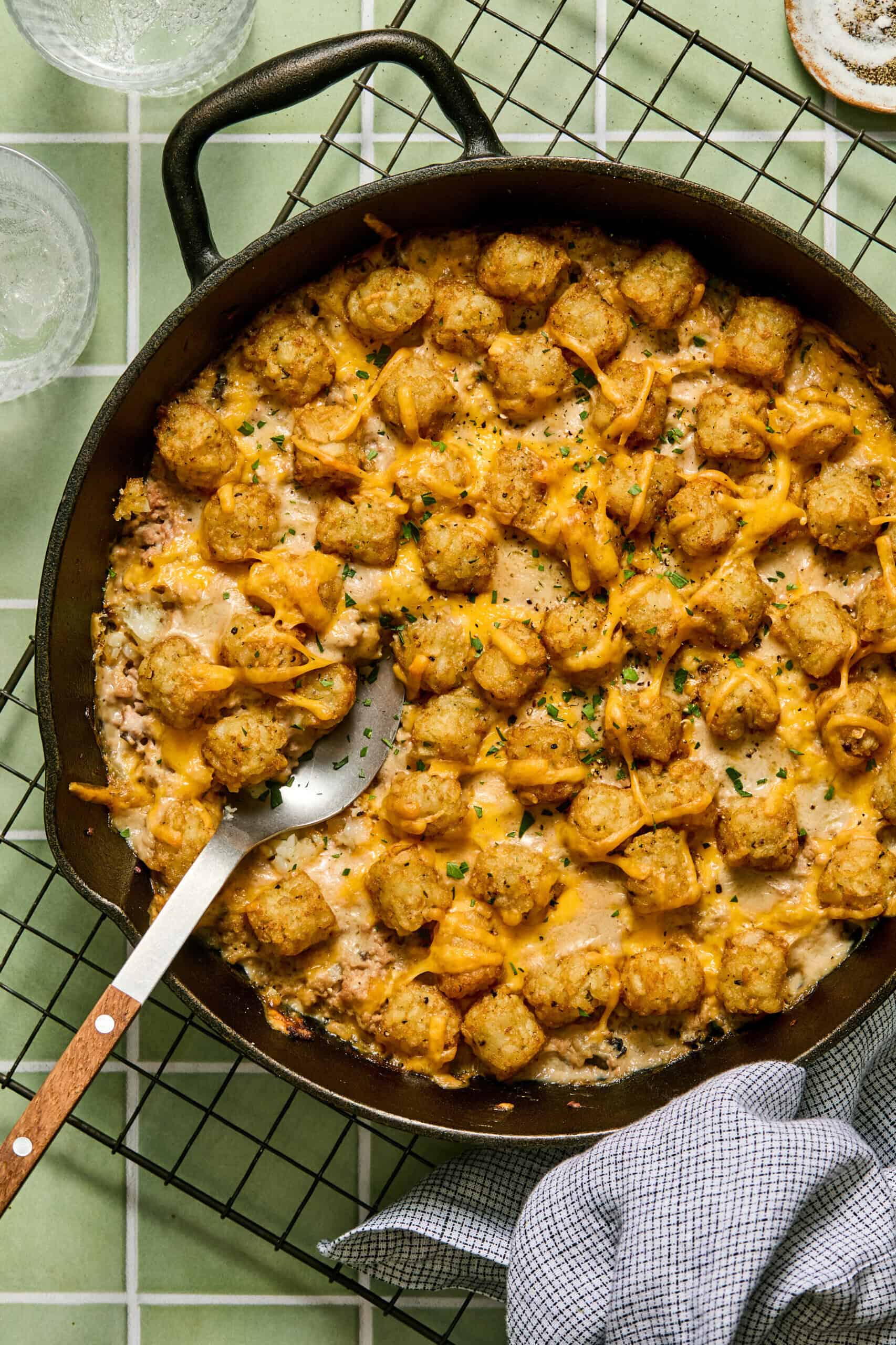 A cast iron skillet filled with a hearty Turkey Tater Tot Casserole, topped with melted cheese and chopped chives. A serving spoon rests in the dish, and a checkered cloth is nearby. The skillet sits on a cooling rack.