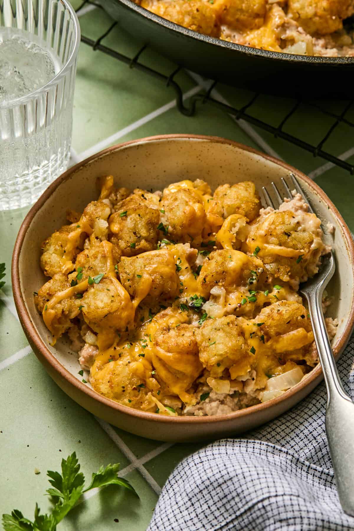 A bowl of Turkey Tater Tot Casserole with melted cheese, ground meat, and parsley, served with a fork on a green tiled table next to a glass of ice water.