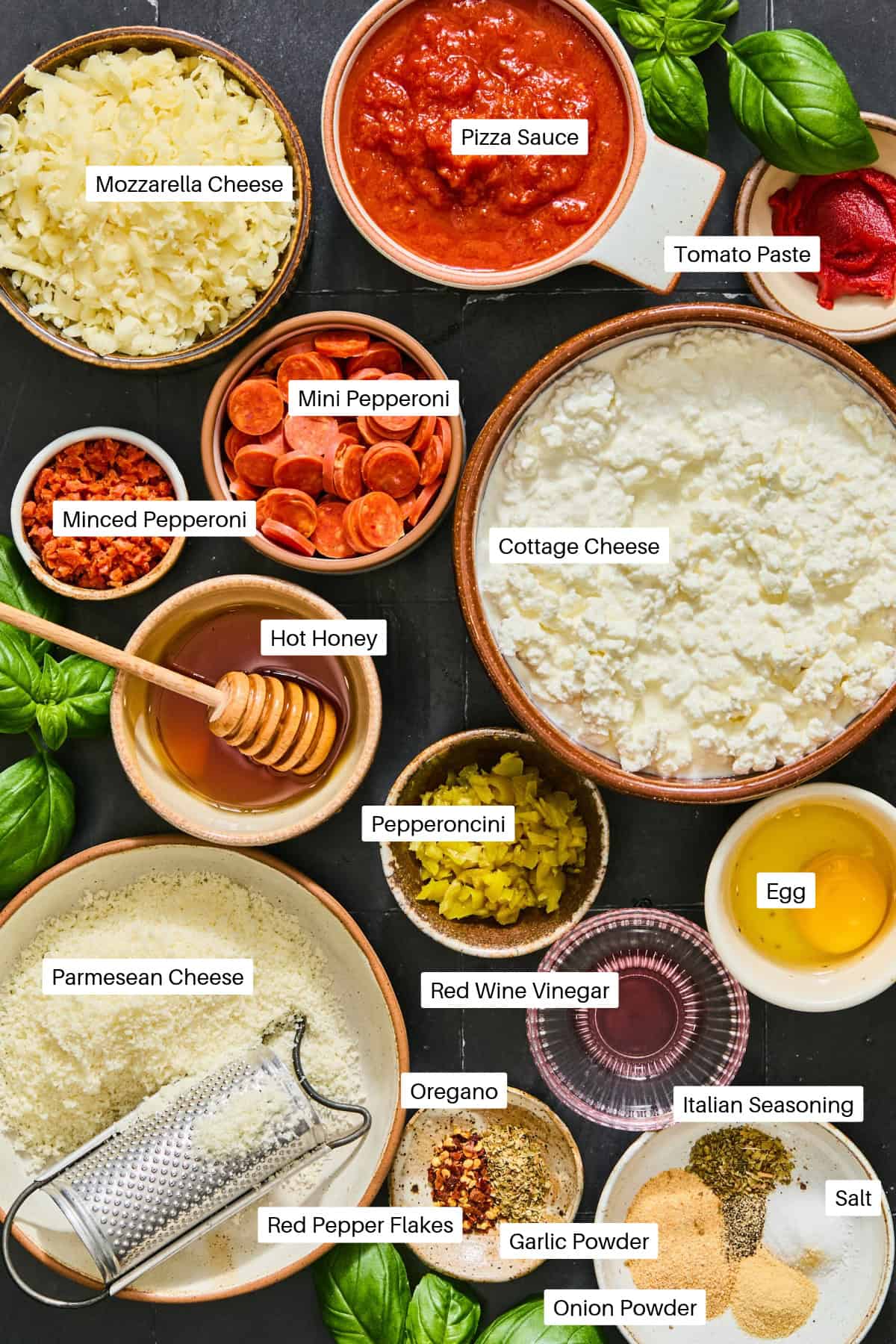 An overhead view of ingredients for a cottage cheese pizza dip, including bowls of mozzarella, pizza sauce, tomato paste, mini and minced pepperoni, cottage cheese, hot honey, pepperoncini, egg, parmesan, red wine vinegar, and various herbs.