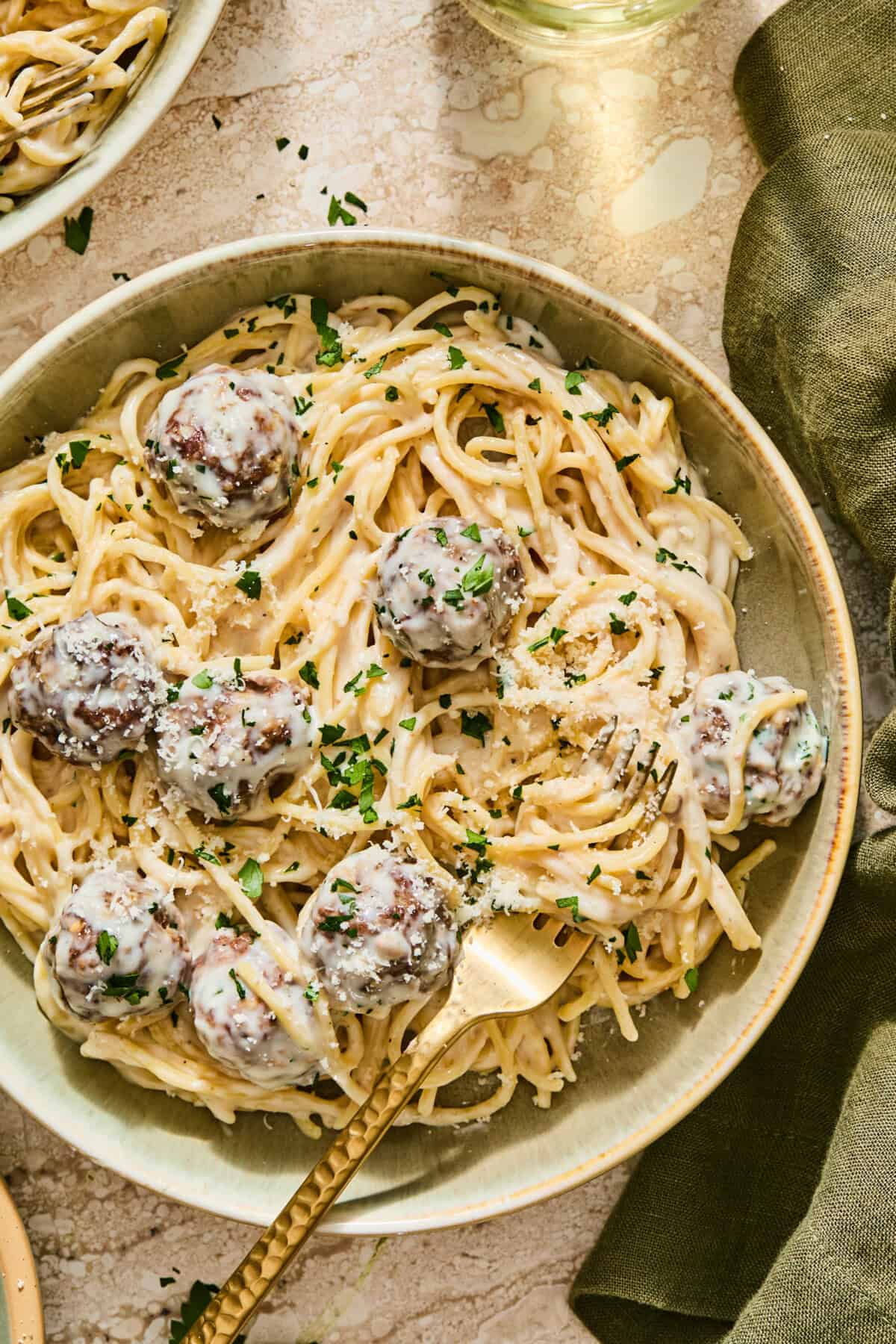 A bowl of Alfredo spaghetti and meatballs in a creamy sauce, garnished with parsley and grated cheese, with a gold fork resting in the dish.
