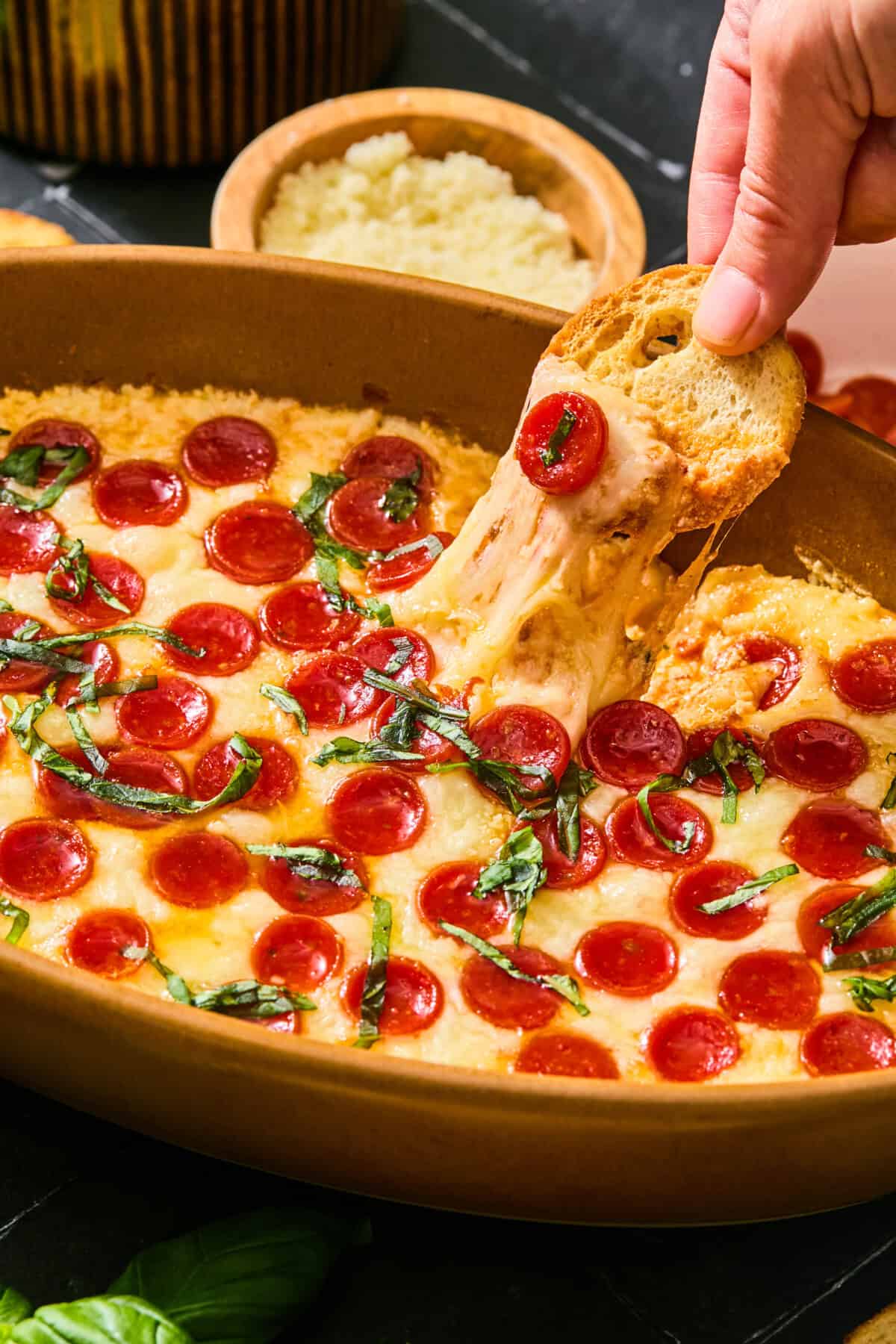 A hand dips a slice of toasted bread into a bubbling cottage cheese pizza dip topped with fresh basil.