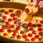 A hand dips a slice of toasted bread into a bubbling cottage cheese pizza dip topped with fresh basil.