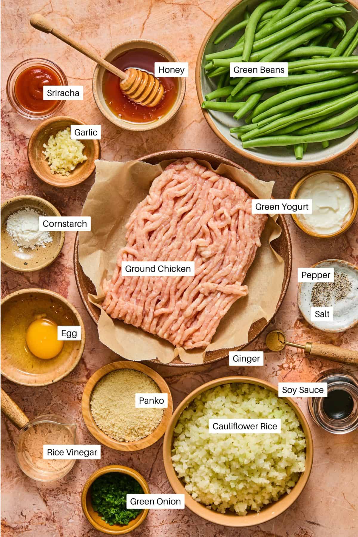 Ingredients for Hot Honey Chicken Meatball Bowls are arranged on a table, including labeled bowls of ground chicken, green beans, cauliflower rice, egg, spices, sauces, Greek yogurt, panko, green onion, and minced garlic and ginger.