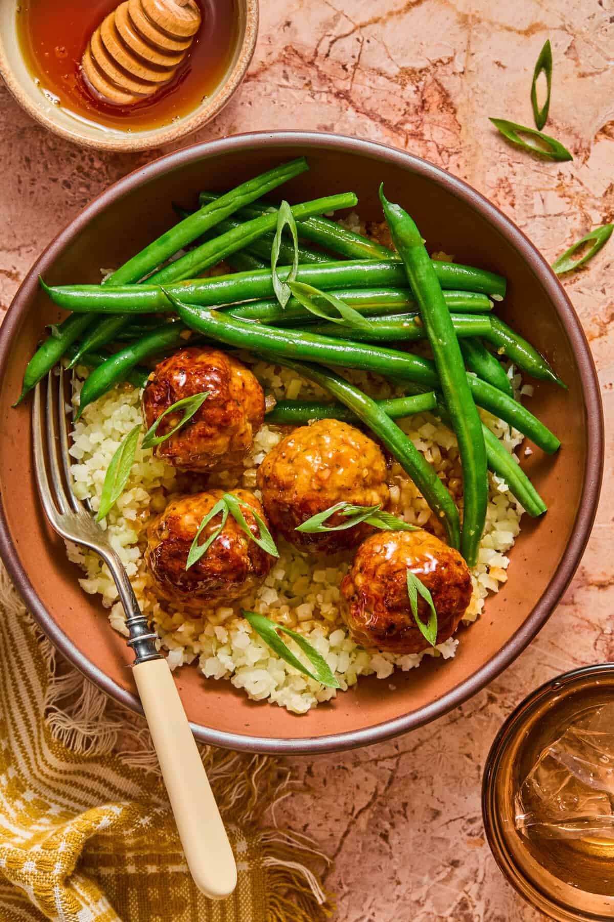 A bowl of cauliflower rice topped with four glazed meatballs, green beans, and sliced green onions, with a fork on the side. This wholesome Hot Honey Chicken Meatball Bowl is served alongside a small bowl of honey and a glass of brown liquid.