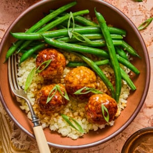 A bowl of cauliflower rice topped with four glazed meatballs, green beans, and sliced green onions, with a fork on the side. This wholesome Hot Honey Chicken Meatball Bowl is served alongside a small bowl of honey and a glass of brown liquid.