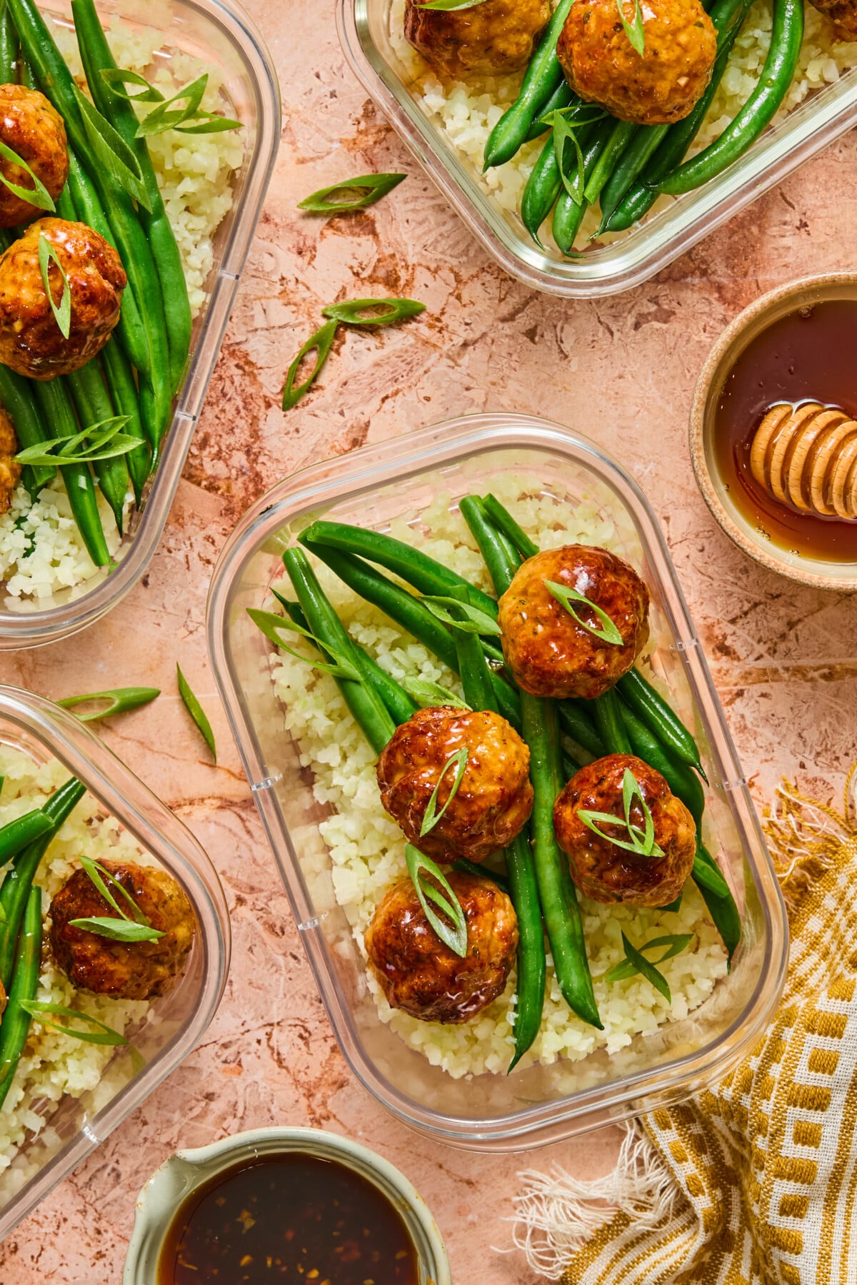 Three meal prep containers with white rice, green beans, and Hot Honey Chicken Meatball Bowls, garnished with sliced green onions. A small bowl of sauce, honey dipper, and striped towel are nearby on a textured peach surface.