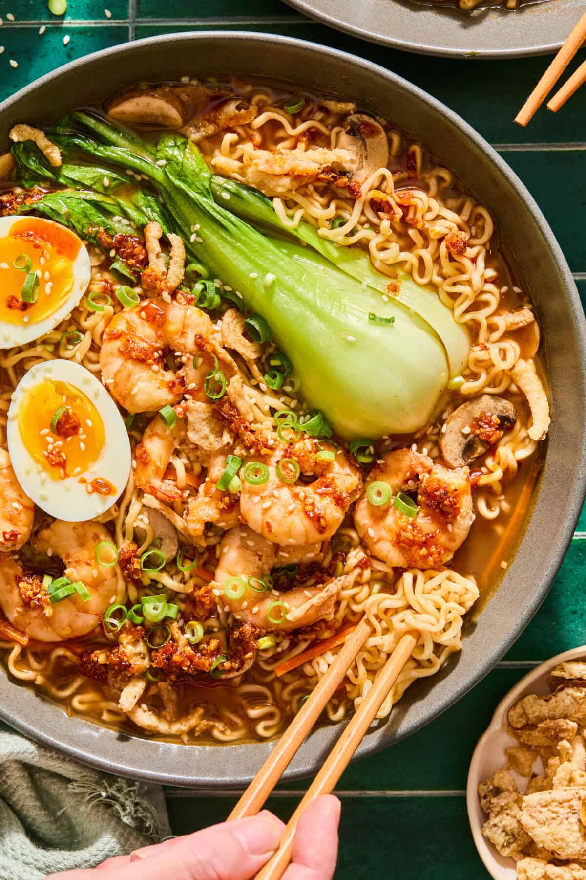 A bowl of Spicy Shrimp Ramen with bok choy, mushrooms, soft-boiled eggs, green onions, and chili oil, with chopsticks picking up noodles.