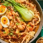 A bowl of Spicy Shrimp Ramen with bok choy, mushrooms, soft-boiled eggs, green onions, and chili oil, with chopsticks picking up noodles.