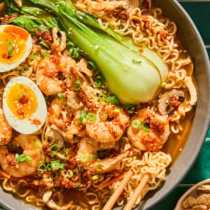 A bowl of Spicy Shrimp Ramen with bok choy, mushrooms, soft-boiled eggs, green onions, and chili oil, with chopsticks picking up noodles.
