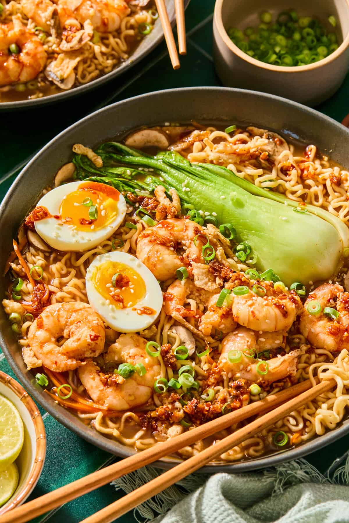 A bowl of Spicy Shrimp Ramen with bok choy, a halved soft-boiled egg, green onions, mushrooms, and chili oil. Chopsticks rest on the bowl, and a small bowl of sliced green onions is nearby.