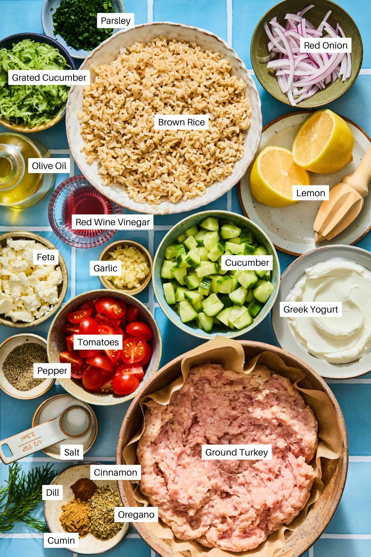 Overhead view of bowls containing ingredients for a turkey gyro bowl meal prep, including ground turkey, brown rice, chopped vegetables, feta, Greek yogurt, and various spices, with labels for each item.