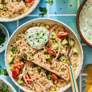 A gyro bowl of rice topped with salmon fillets, cucumber tomato salad, and a dollop of tzatziki sauce.