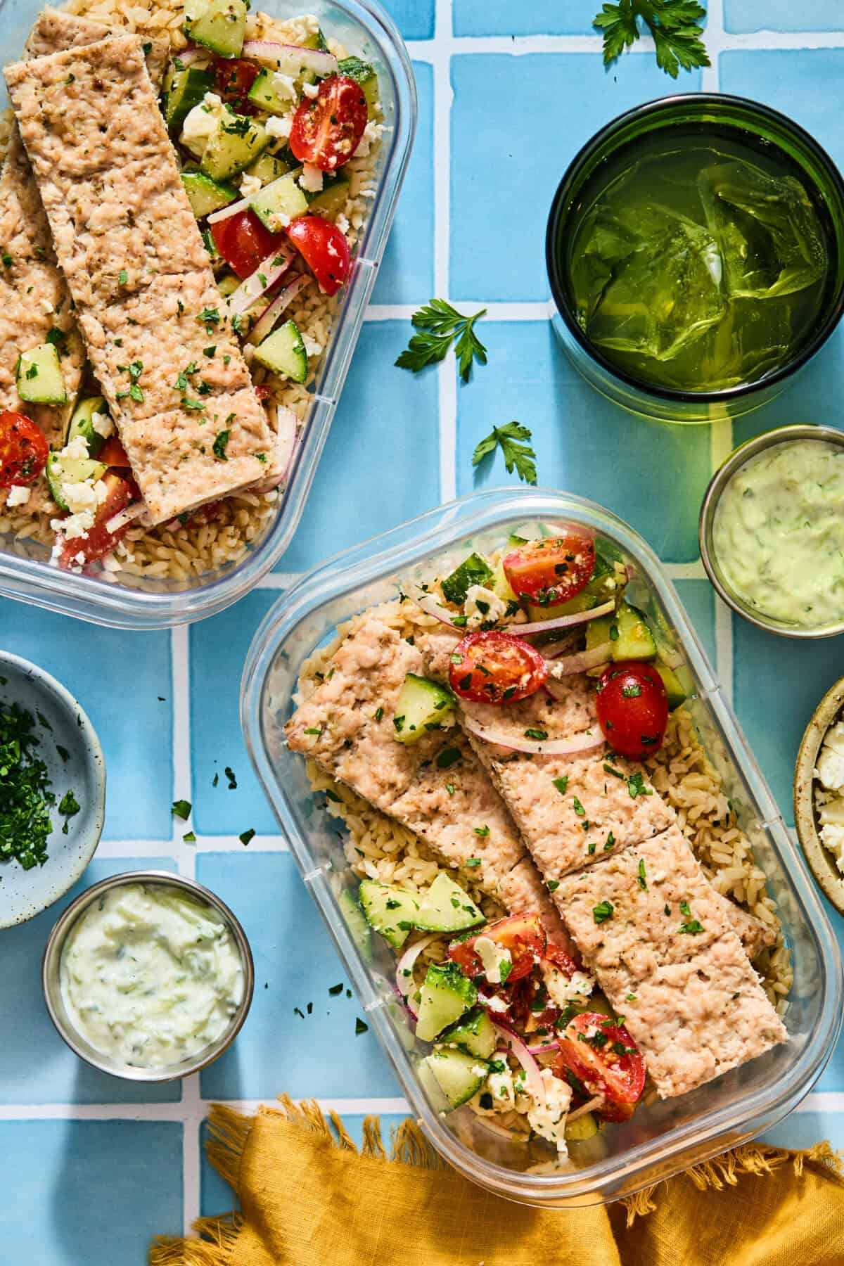 Two plastic gyro bowls filled with rice, salmon fillets, and a cucumber-tomato salad, served with small bowls of tzatziki sauce.
