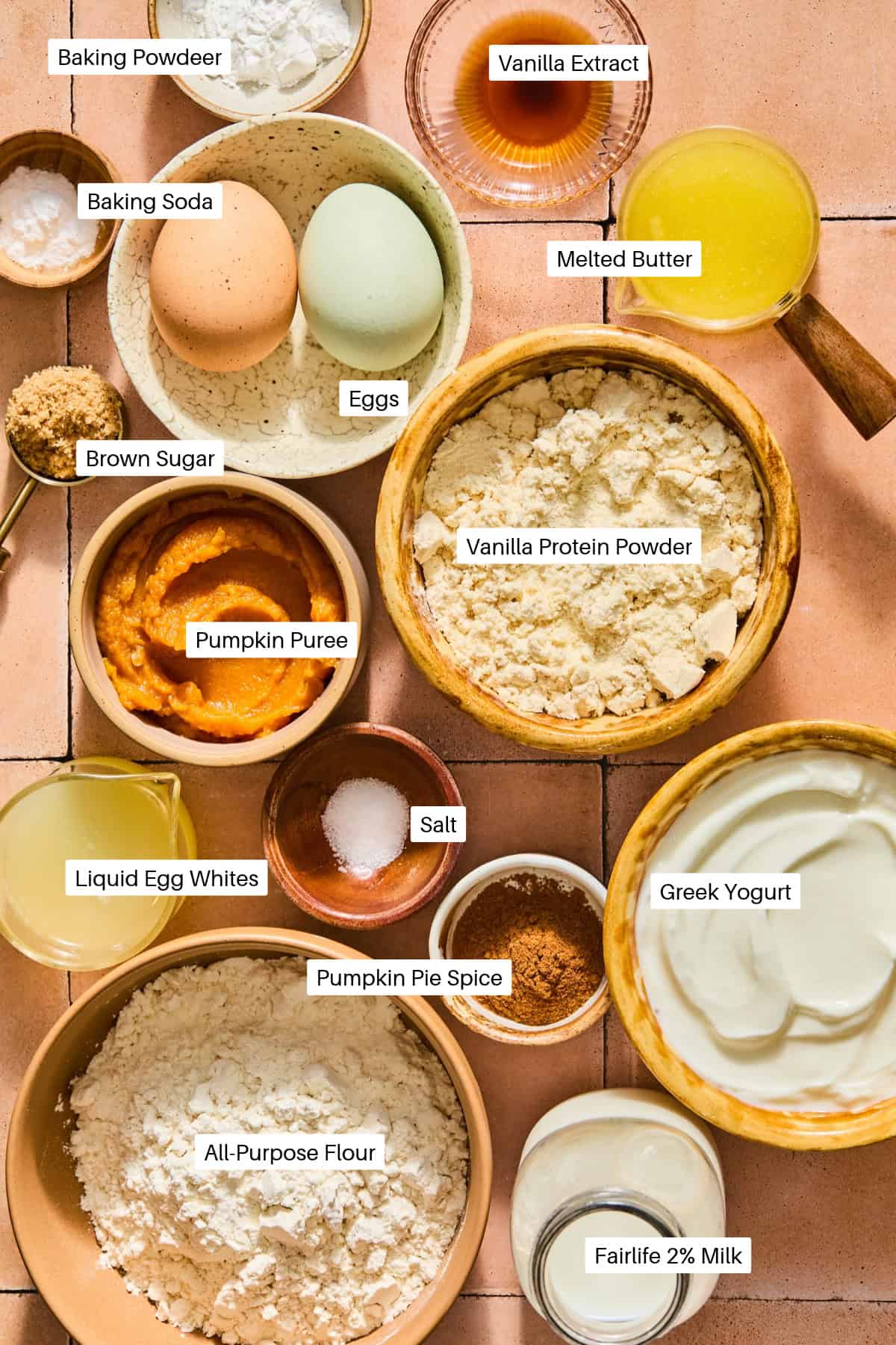 Ingredients for pumpkin protein muffins and pumpkin pancakes in small bowls, labeled: baking powder, vanilla extract, baking soda, eggs, melted butter, brown sugar, pumpkin puree, protein powder, egg whites, salt, pumpkin pie spice, yogurt, and flour.