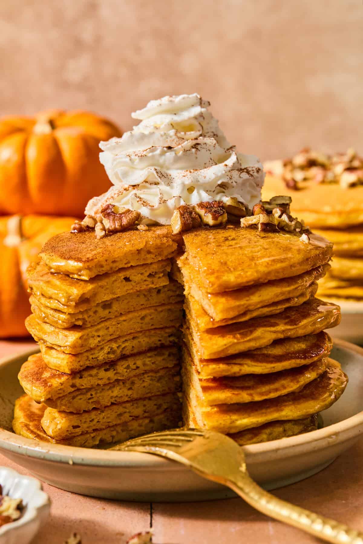 A stack of pumpkin pancakes topped with whipped cream and pecans, with a wedge cut out to reveal the interior texture.