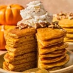 A stack of pumpkin pancakes topped with whipped cream and pecans, with a wedge cut out to reveal the interior texture.