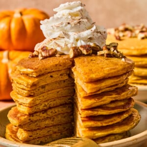 A stack of pumpkin pancakes topped with whipped cream and pecans, with a wedge cut out to reveal the interior texture.
